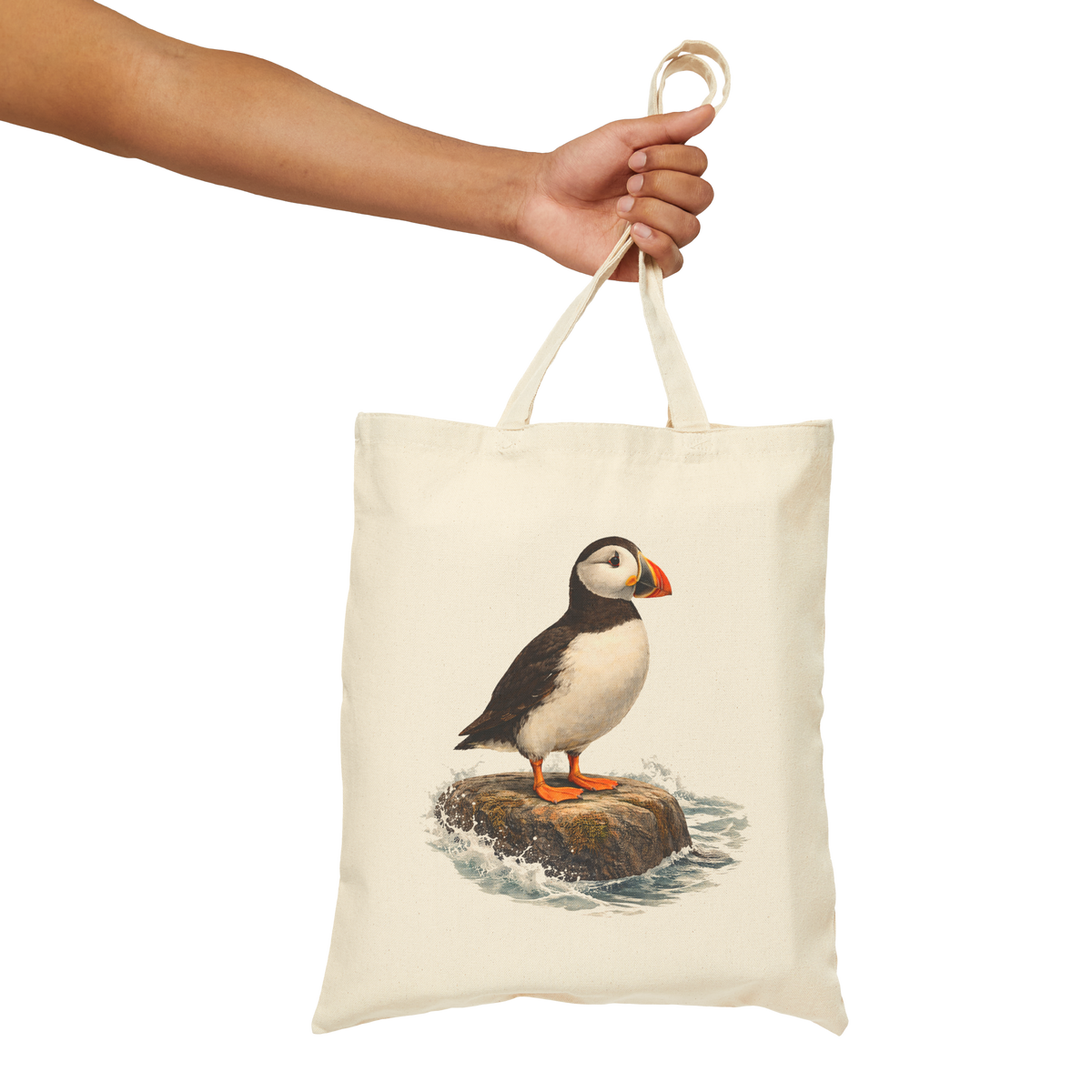 Natural cotton canvas tote bag held by its handles, printed with a puffin standing on a coastal rock