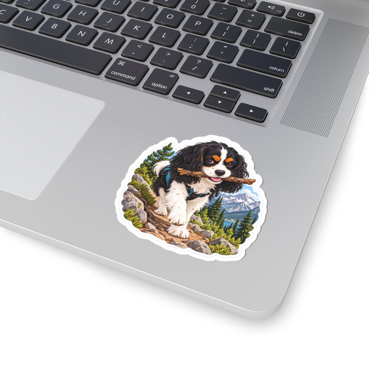 Product mockup of the Cavy Play Mode Trail Stick kiss-cut sticker: a tricolor Cavalier King Charles Spaniel carrying a stick along a mountain trail with pines and snowy peaks.