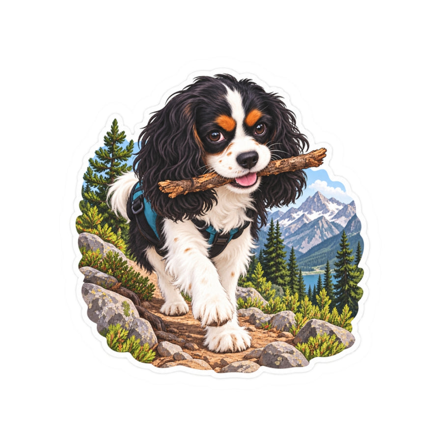 Product mockup of the Cavy Play Mode Trail Stick kiss-cut sticker: a tricolor Cavalier King Charles Spaniel carrying a stick along a mountain trail with pines and snowy peaks.